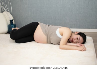 A Japanese Asian young beautiful pregnant woman lying on her side with relaxed clothes pulls up her clothes exercises regime to convert breech presentation on a white carpet with blue background from 