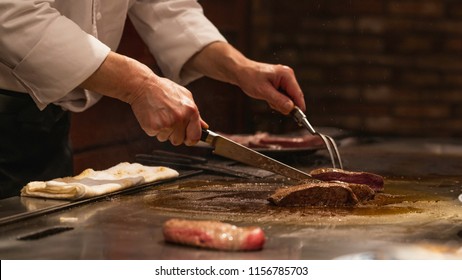 A Japanese chef hand holding a knife and fox is cooking a sliced Kobe Beef or Wagyu steak in pan in Teppanyaki style. It is hot and delicious. 
