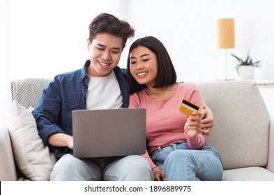 Japanese Couple Shopping Online Using Laptop And Credit Card Paying For Order Sitting On Couch At Home. Family Buying And Purchasing Things On Internet. Weekend Shopping Concept