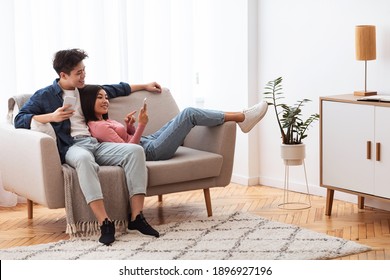 Japanese Couple Using Cellphones Texting And Browsing Internet Relaxing Sitting On Couch Indoor. Weekend Leisure. Happy And Relaxed Asian Family Using Mobile Application Resting At Home