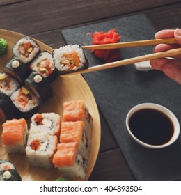 Japanese food restaurant, maki plate, platter set. Hand takes roll. Set with chopsticks, ginger, soy sauce at rustic wood background and black stone. POV at black. Instagram style. 