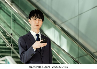 A Japanese man in his 20s in a suit with a camera looking at a camera with a smartphone while traveling on an escalator