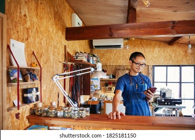 Japanese Man Wearing Blue Apron And Glasses Standing In A Leather Shop, Using His Mobile Phone.