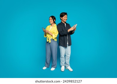 A japanese millennial man and woman stand back to back, each engrossed in their own smartphones, dressed in casual clothing with pleasant expressions against a bright blue background - Powered by Shutterstock