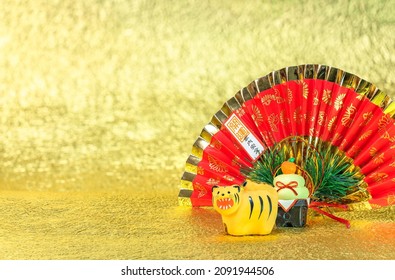 Japanese New Year's Greeting Card Depicting A Zodiac Animal Figurine Of A Roaring Tiger For 2022 With A Folding Fan Written Good Luck For A Safe House And A Kagami Mochi Against A Gold Background.