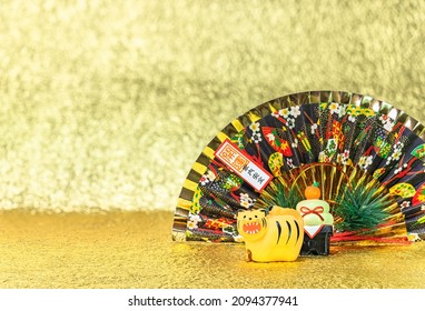 Japanese New Year's Greeting Card Of A Funny Roaring Tiger Figurine For The Year 2022 With A Folding Fan Written Good Luck For A Safe House Against A Gold Background.