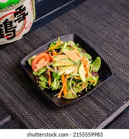 Japanese salad with tomato, cucumber and sesame dressing in the traditional Japanese ramen restaurant, with a black plate on a black mat and Japanese fonts sake barrel translation: Snow Crane