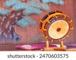 Japanese shime-daiko drum whose edges are laced with thick orange rope placed on a wooden terendai support with bachi sticks next to a zabuton cushion in front of a giant mural depicting a pine tree.