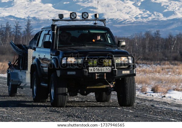 Japanese Sport Utility Vehicle Toyota Land Cruiser\
with trailer which carries snowmobile driving along road on\
background of winter landscape sunny day. Kamchatka Peninsula,\
Russia - January 7,\
2016