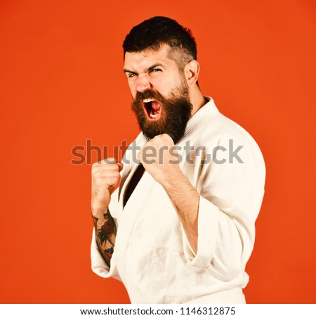 Japanese sports and martial arts concept. Young sporty man in white kimono isolated on red background. Athlete with beard and raging face holds fists in defence.