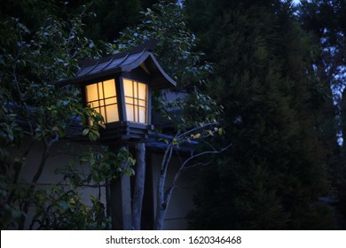 Japanese Style Garden Lamp. Japanese Gable Roof House Light Post In Traditional Garden. Warm Light In Cool Sunset Garden. 