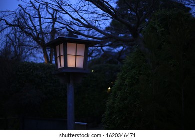 Japanese Style Garden Lamp. Japanese Gable Roof House Light Post In Traditional Garden. Warm Light In Cool Sunset Garden. 