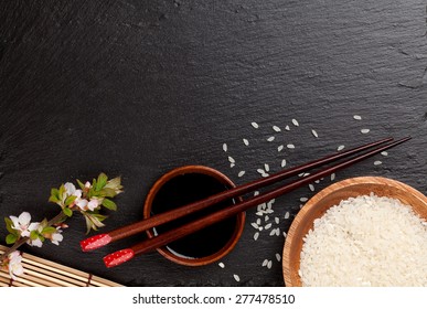 Japanese sushi chopsticks over soy sauce bowl, rice and sakura blossom on black stone background. Top view with copy space