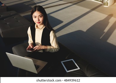 Japanese Woman CEO Is Using Mobile Phone And Laptop Computer Before Conference With Staff. Female Skilled Economist With Cellphone In Hands Is Posing During Work On Net-book In Co-working Space