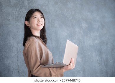 Mulher japonesa com computador no fundo cinza escuro. Ela olha para cima com um sorriso. Copie o espaço à direita.