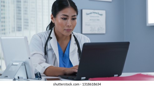 Japanese Woman Doctor Sitting At Desk Working On Laptop Computer. Female Primary Care Physician Making Notes On Patient Records Using Computer