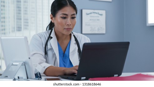 Japanese Woman Doctor Sitting At Desk Working On Laptop Computer. Female Primary Care Physician Making Notes On Patient Records Using Computer