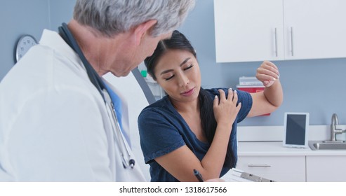 Japanese Woman Explaining Shoulder Pain To Doctor In Exam Room. Patient With Rotator Cuff Injury Talking To Medical Professional