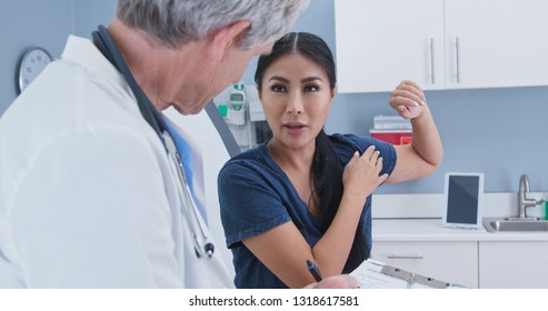 Japanese Woman Explaining Shoulder Pain To Doctor In Exam Room. Patient With Rotator Cuff Injury Talking To Medical Professional