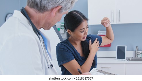 Japanese Woman Explaining Shoulder Pain To Doctor In Exam Room. Patient With Rotator Cuff Injury Talking To Medical Professional