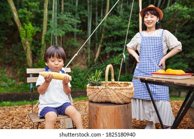 Japanese Woman Outdoors And Young Boy Sitting On Chair, Eating Corn On The Cob.