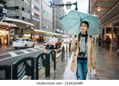 japanese woman pedestrian with camera and plastic bag is walking on sidewalk when it's drizzling. asian female traveler carrying camera is strolling back to hotel after she's done grocery shopping. - Powered by Shutterstock