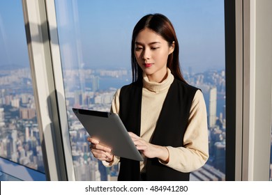 Japanese Woman Professional Worker In Real Estate Company Is Reading Advertising In Internet Via Digital Tablet, While Is Standing In Modern Office Interior Near Window With View Of Business District