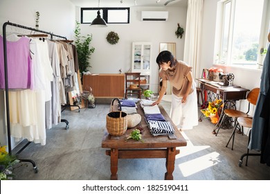 Japanese woman standing in a small fashion boutique, looking at T-Shirts on a coffee table. - Shutterstock ID 1825190411