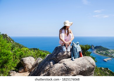 Japanese woman wearing hat sitting on rock on a cliff, ocean in the background. - Shutterstock ID 1825176068