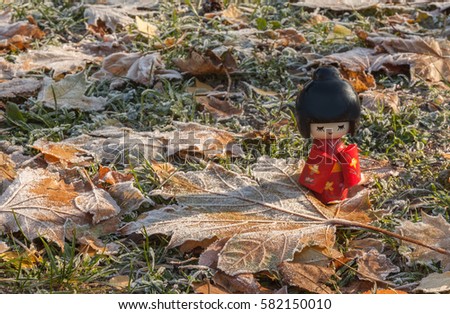 Japanese wooden doll Kokeshi on the background of maple leaves with frost. Concept season maple leaves Momiji-gari. (Mass-produced products)