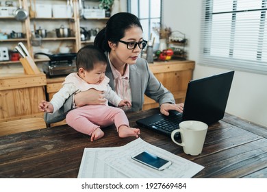 japanese working mother is typing on laptop single handed and putting her lovely kid on table. asian female worker letting her cute baby sitting on desk is teleworking with notebook at home. - Powered by Shutterstock