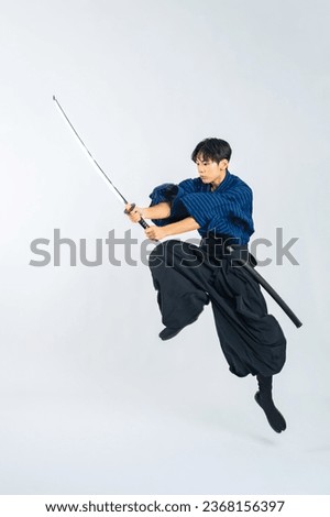 Japanese young samurai wielding katana sword while jumping in studio with white background.