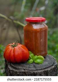 Jar With Adjika On The Background Of A Garden Stands On A Stump. Fresh Red Ribbed Tomato, Basil With A Jar Of Ketchup For Sale At The Farmers Market. Concept Of Gardening And Delivery Of Organic Food