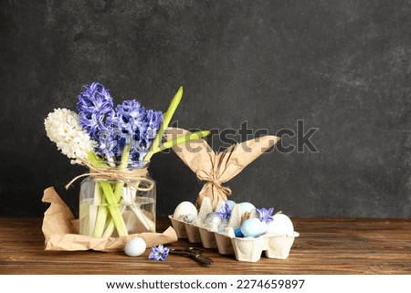 Jar with beautiful hyacinth flowers and Easter eggs in package on wooden table near black wall