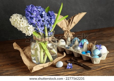 Jar with beautiful hyacinth flowers and Easter eggs in package on wooden table near black wall
