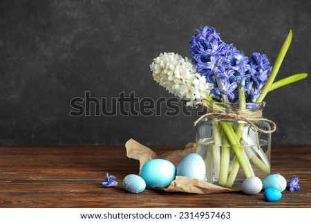 Jar with beautiful hyacinth flowers and Easter eggs on wooden table near black wall