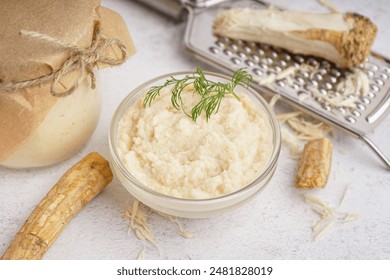 Jar and bowl of horseradish sauce with horseradish roots on white background