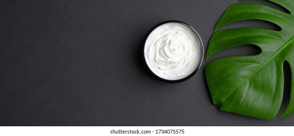 Jar With Cosmetic Cream And Tropical Monstera Leaf On Black Background. Natural Organic Beauty Product For Body, Hand, Face Skin Treatment. Flat Lay, Top View. Skincare Concept.