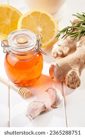 Jar of honey by some ginger, a clove of garlic, rosemary leaves and a sliced yellow lemon over a white wooden table. Natural remedies. - Shutterstock ID 2365288751