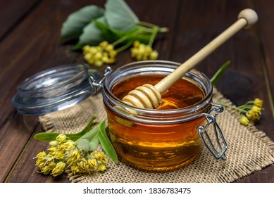 Jar with linden honey with stick for honey and fresh linden flowers on a wooden table.