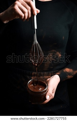 A jar of melted chocolate in the hands of a chocolatier. Kitchen whisk. On a black background.