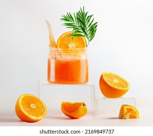 Jar Of Orange Facial Peeling Mask At Podium. Spa Setting With Orange Sugar Scrub With Tropical Green Leaf, Wooden Massage Brush And Orange Fruits. Healthy Skin Care And Beauty Treatment. Front View.