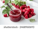 Jar of raspberry jam and fresh berries with leaves on a white wooden table.