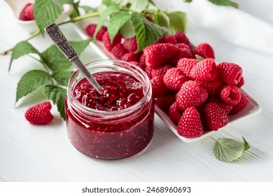 Jar of raspberry jam and fresh berries with leaves on a white wooden table.