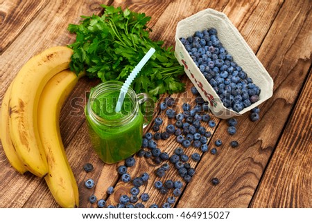 Jar tumbler mug with green smoothie drink and a bundle of fresh parsley, banana and blueberries on wooden table