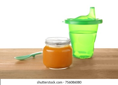 Jar With Yummy Baby Food And Sippy Cup On Wooden Table Against White Background