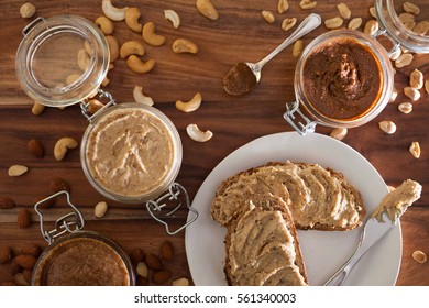 Jars of almond, cashew, and peanut butters sitting in sequence with their respective nuts lying around them
