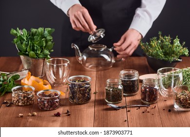 Jars With Assorted Tea, Orange Peel, Fresh Mint, Thyme And Rosemary On Wooden Table. Tea Master Preparing A Glass Pot To Brew Some Tea.