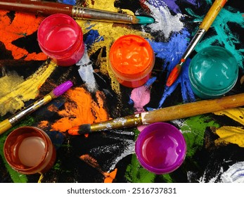 Jars with multi-colored paints, brushes are on a black cloth with spots of different shapes and colors from gouache and acrylic paints, background - Powered by Shutterstock