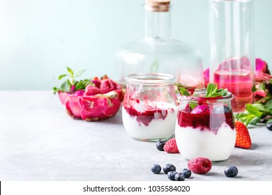 Jars of natural white yogurt with berry sauce, fruit salad with pink dragon fruit, berries and mint, served with bottle of lemonade on grey table. Healthy eating. Copy space